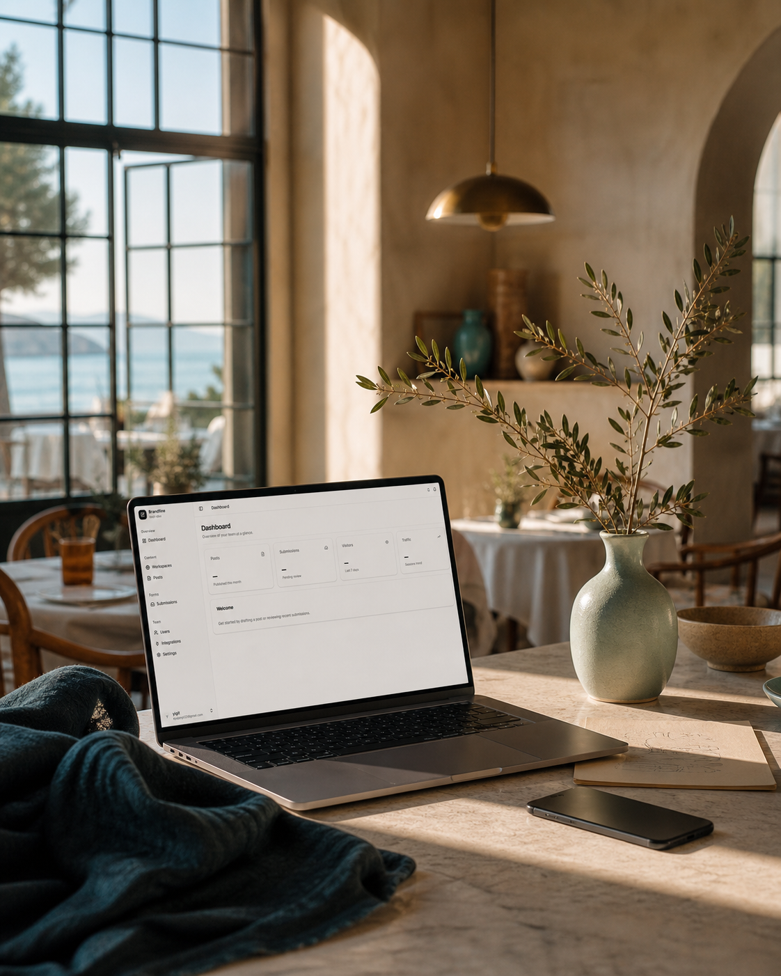 Sunlit Mediterranean restaurant interior with a sea view, an open laptop on a stone table, and olive branches in a ceramic vase.